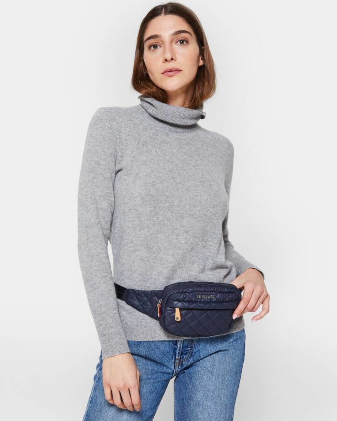 Model wearing a Mz Wallace Metro Belt bag also used as a sling bag in color dawn wearing blue jeans and a gray turtle neck on a white background 