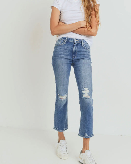 Model wearing just black denim the official weekend jean with rips in the knees wearing white shoes and a white tee shirt crossing her arms on a white background