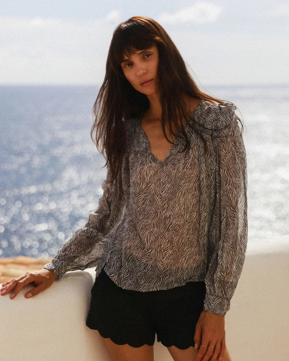 Woman in a patterned blouse and black shorts standing by the ocean.