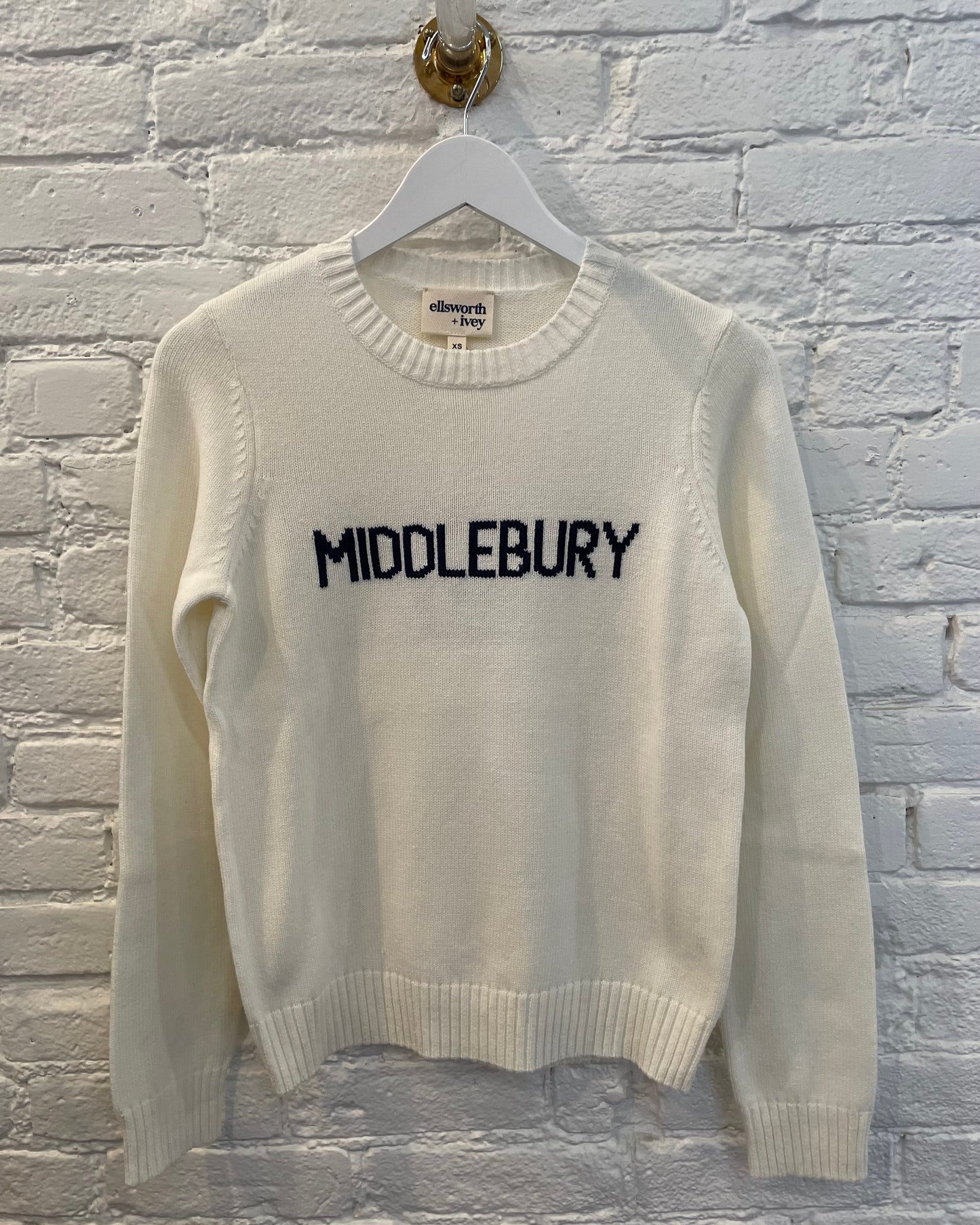White sweater with 'MIDDLEBURY' text on a hanger against a brick wall.