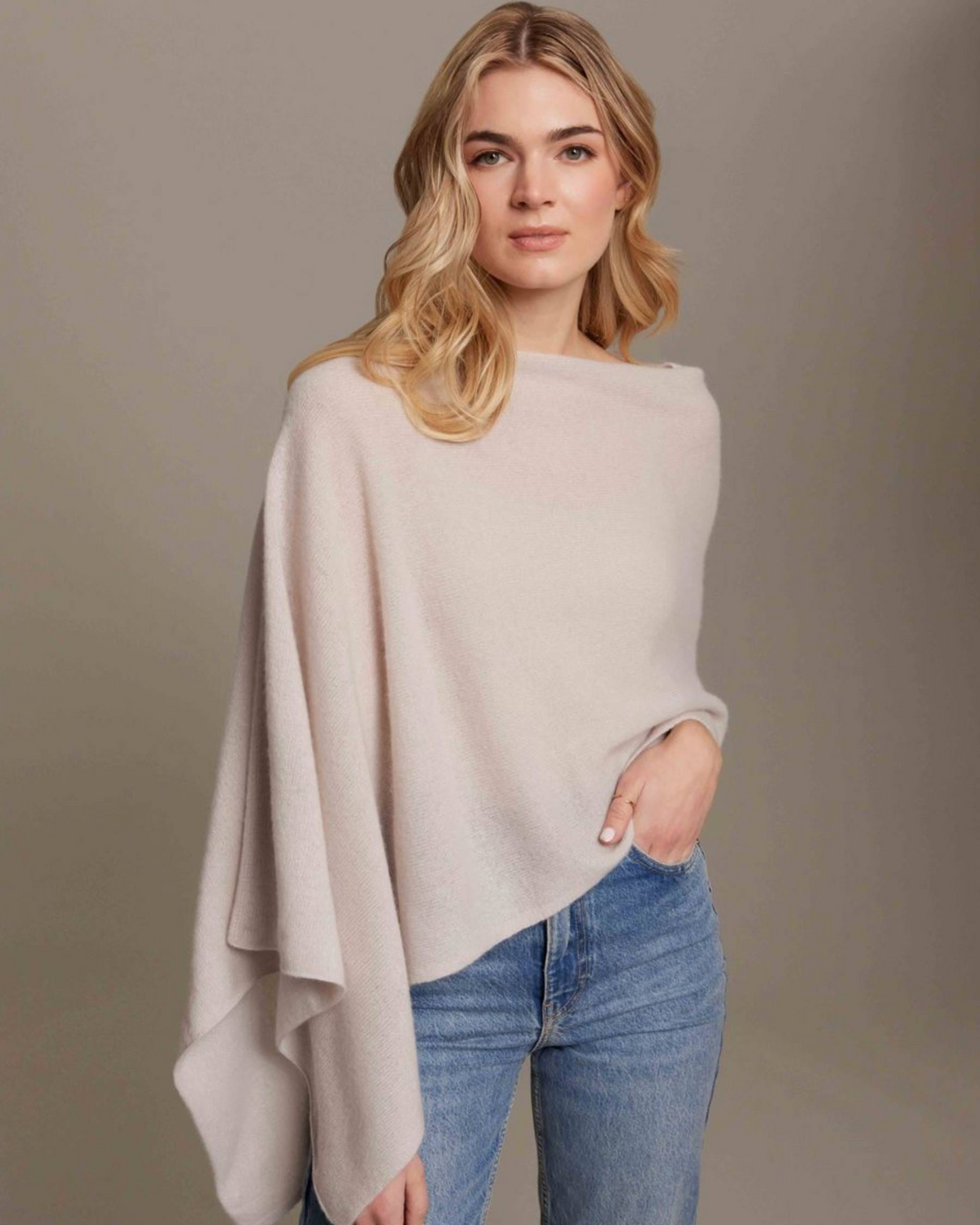 Woman wearing a light beige poncho over a white top and blue jeans on a plain background