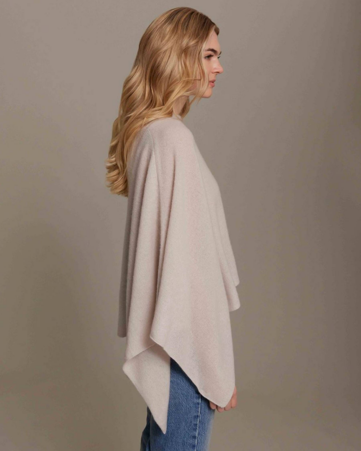 A woman in profile wearing a light beige draped poncho sweater over blue jeans, photographed against a solid neutral gray studio background.