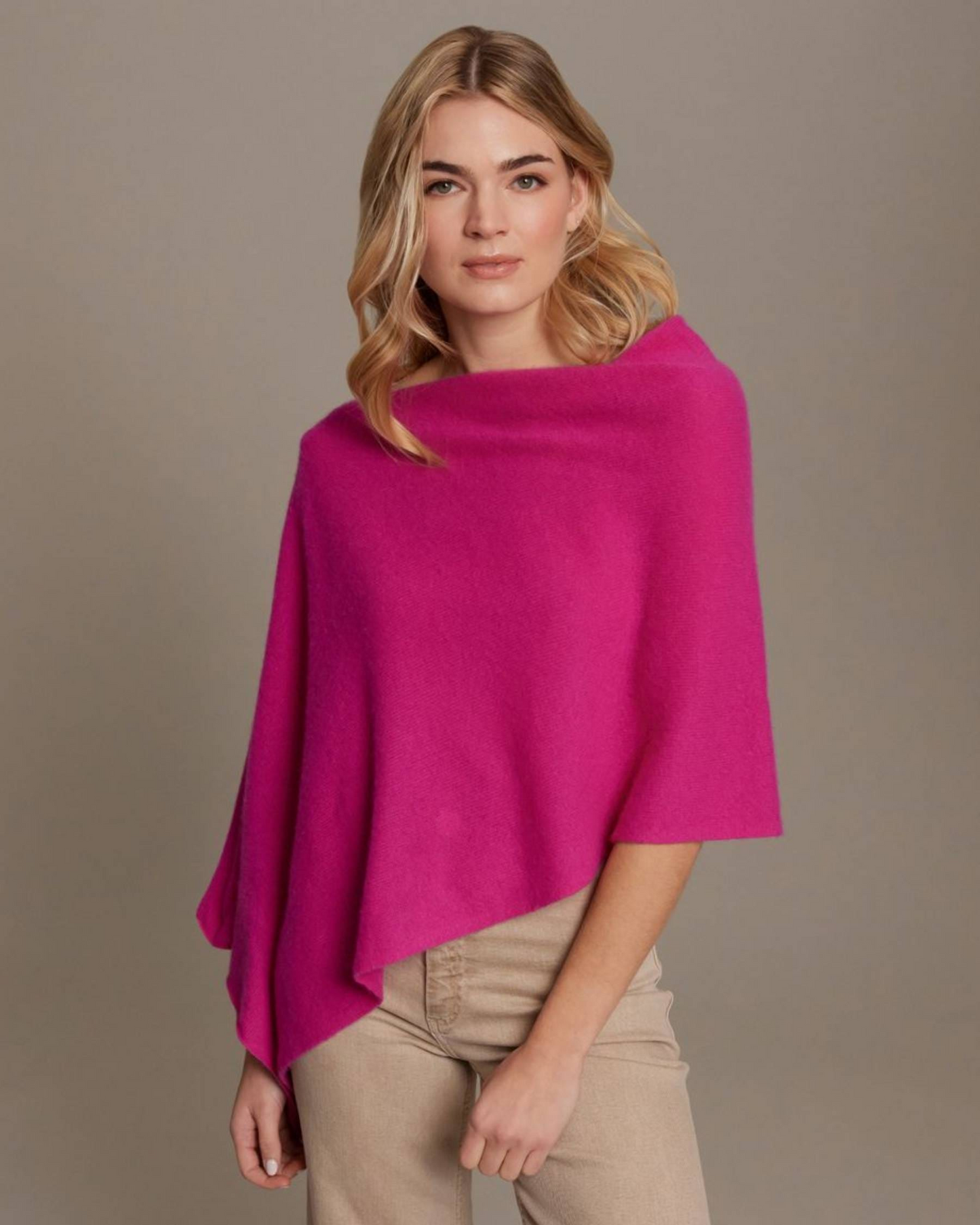A woman modeling a bright magenta knit poncho over beige pants, photographed against a plain warm gray studio background.