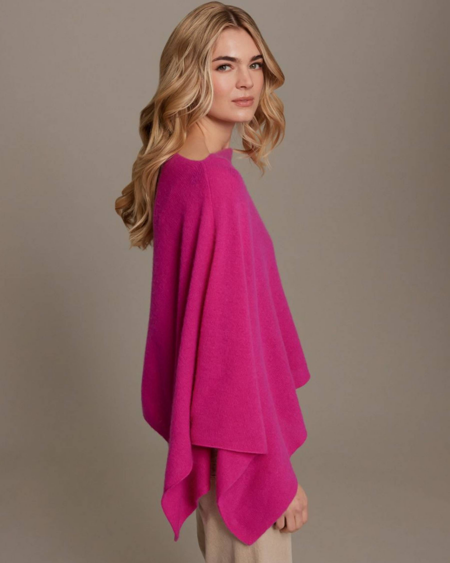A woman with blonde wavy hair is shown from the side wearing a bright pink knit poncho over light beige pants, set against a solid warm gray studio background.