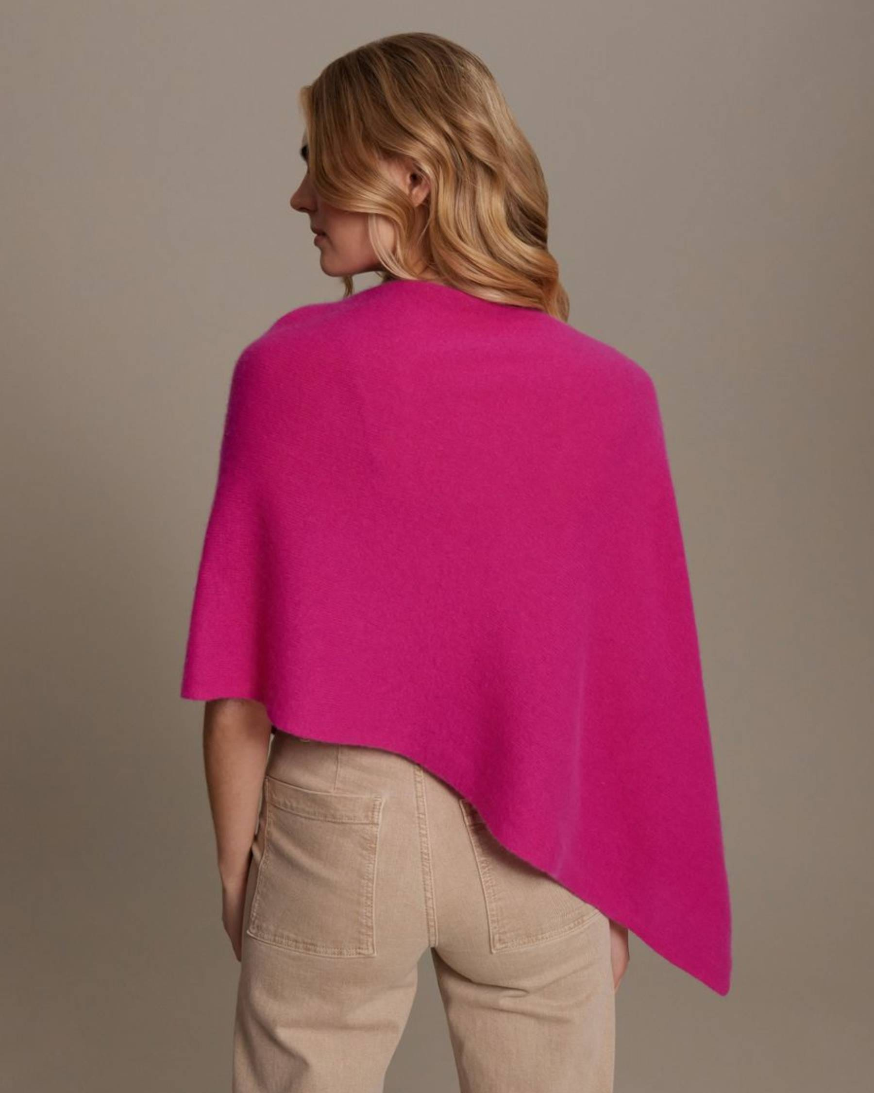 A woman shown from the back wearing a bright pink open-front shawl or poncho over a cream top and beige trousers, photographed against a plain warm gray studio background.