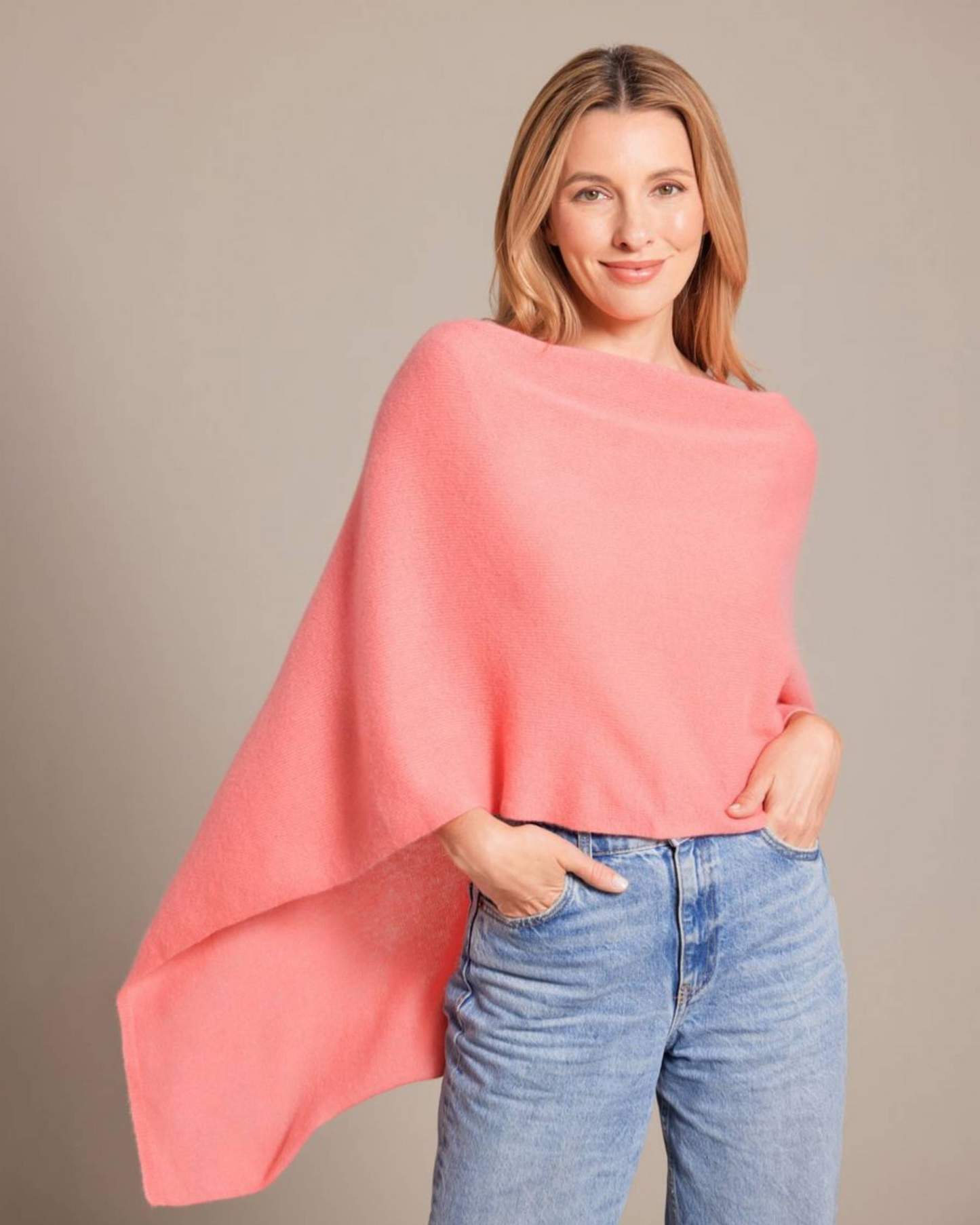 A woman wearing a soft coral pink knit poncho sweater and light blue jeans, standing against a solid neutral beige background.