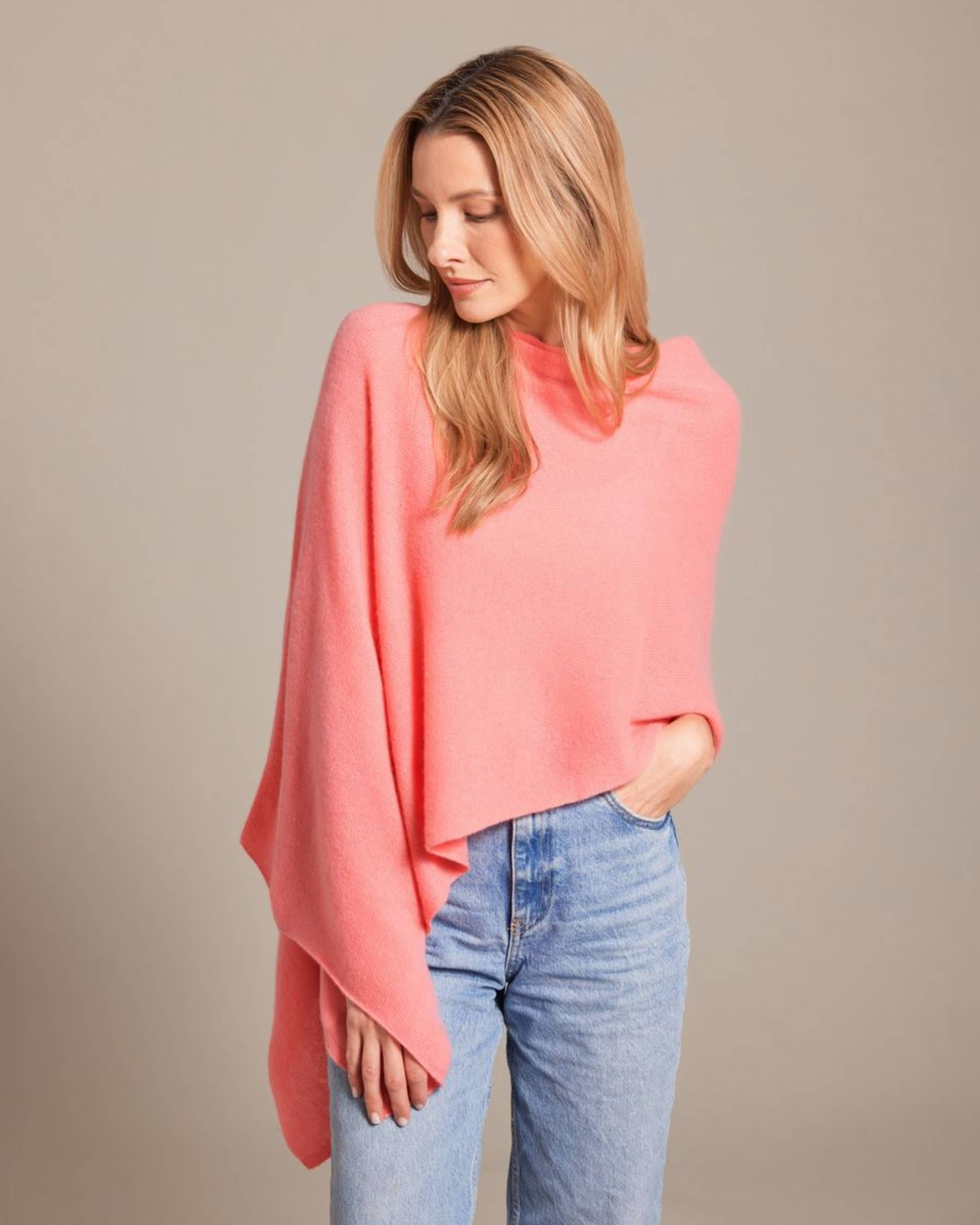 A woman wearing a coral pink knit poncho sweater and light wash blue jeans, standing against a neutral beige studio background.