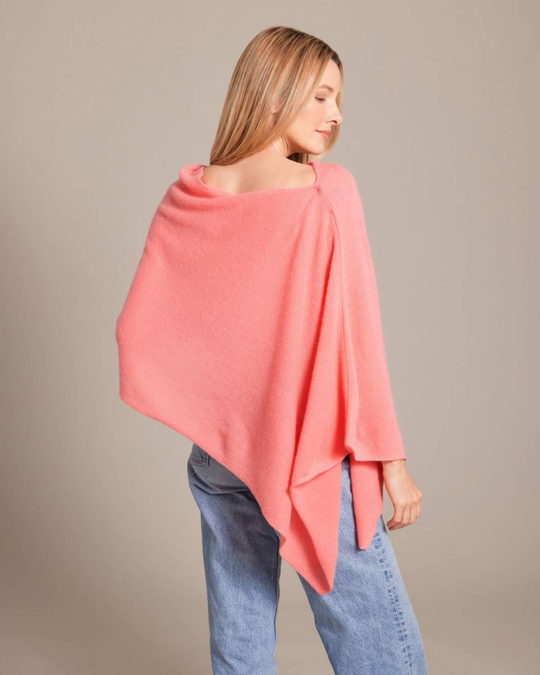 A woman wearing a coral pink knit poncho sweater and light blue jeans, photographed from the back against a solid beige studio background.