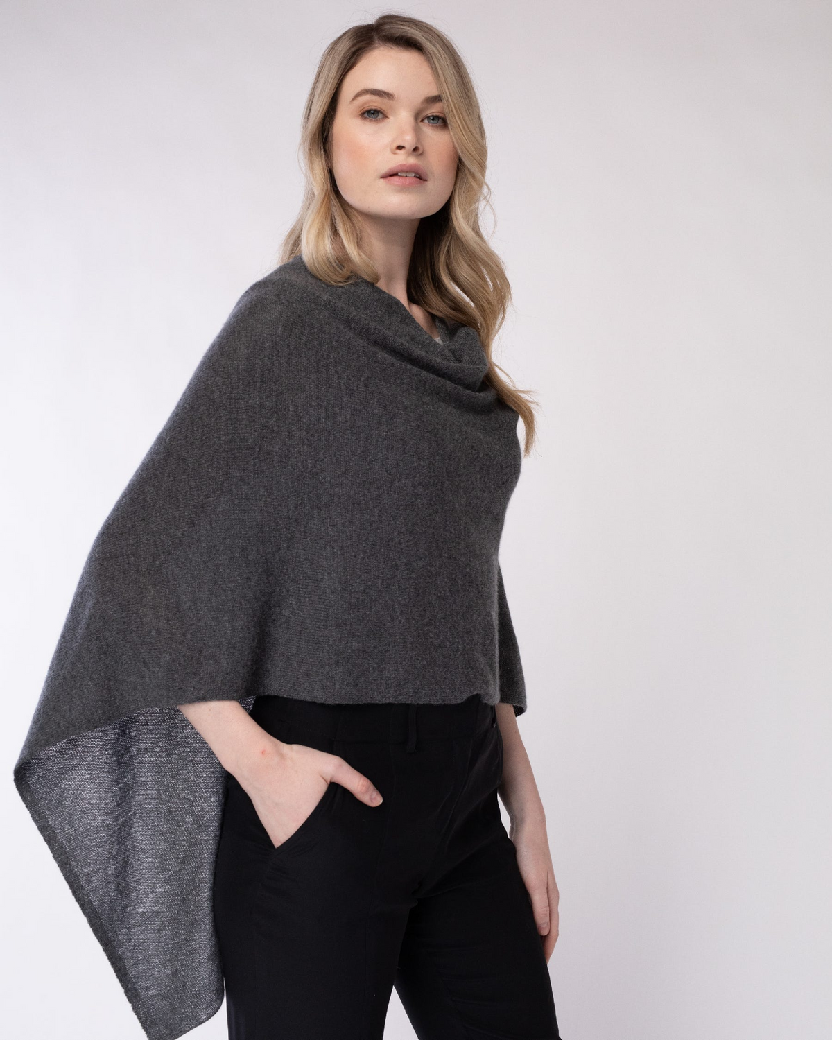 A woman modeling a dark gray knit cowl-neck poncho shawl, paired with black pants, against a plain light gray studio background.