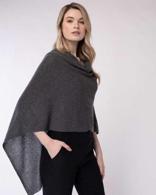 A woman modeling a dark gray knit cowl-neck poncho shawl, paired with black pants, against a plain light gray studio background.