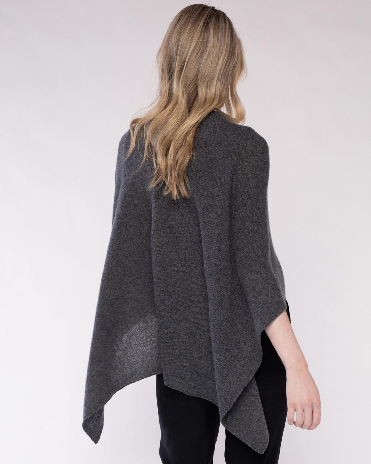 A woman viewed from behind wearing a dark gray open-front poncho or cape over black pants, against a plain light gray studio background.