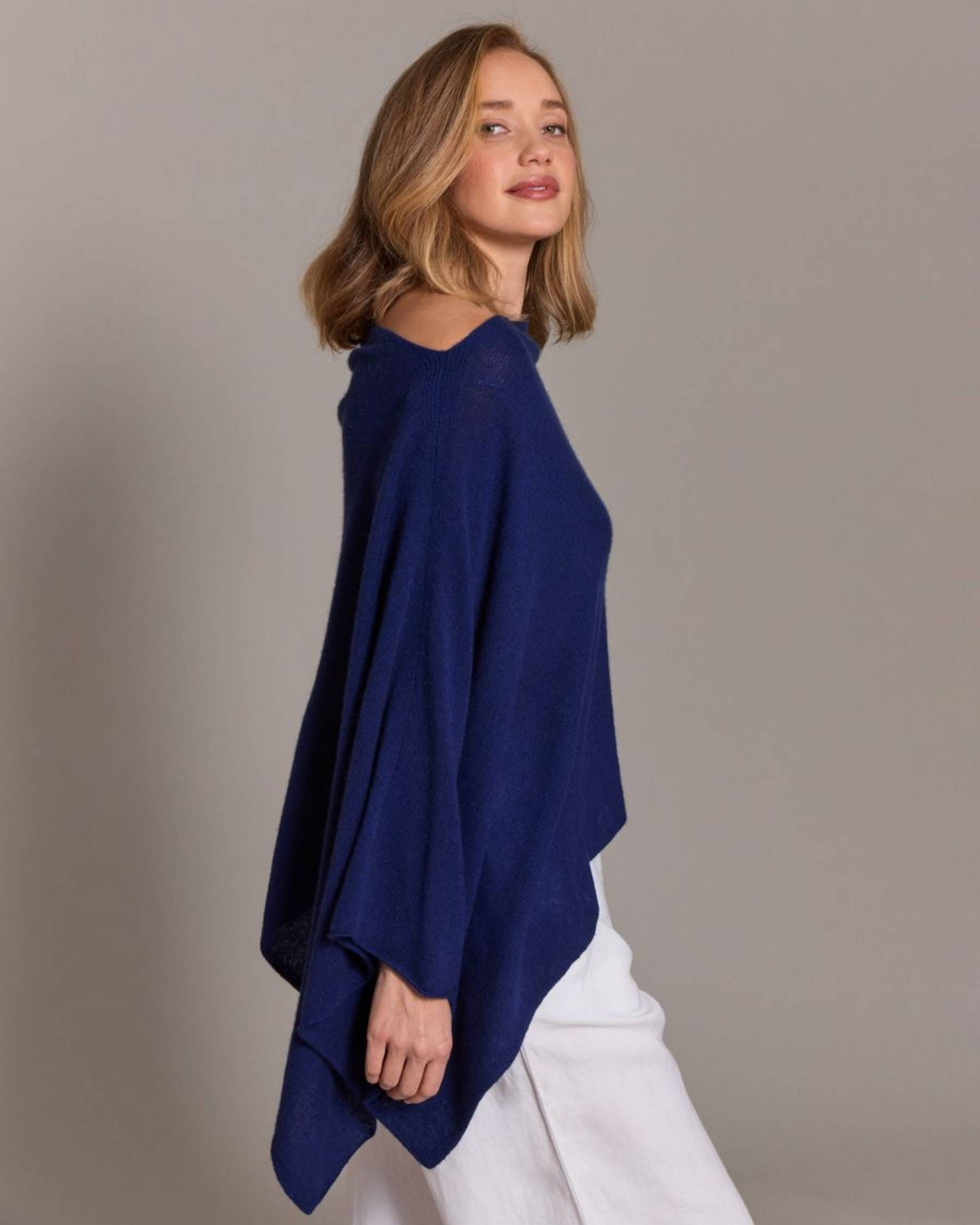 A woman wearing a navy blue knit poncho over white pants, photographed against a plain light gray studio background.
