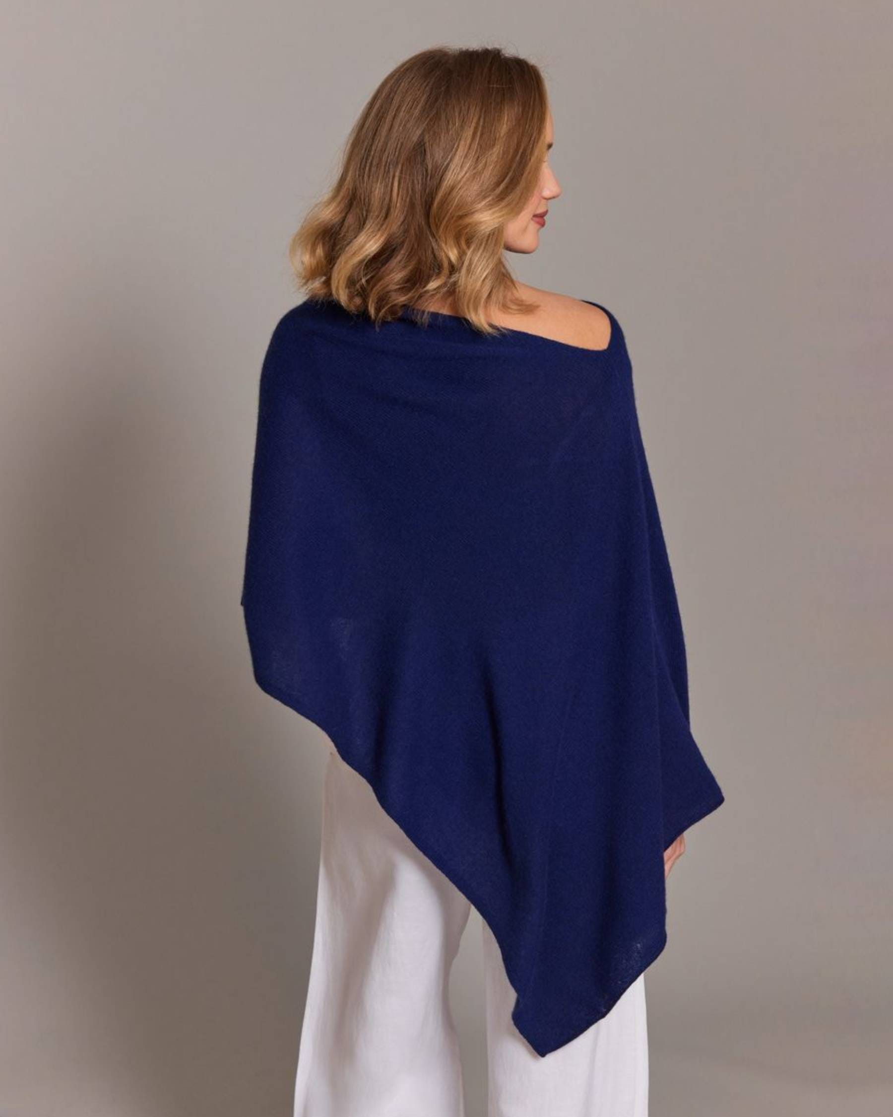 A woman shown from the back wearing a navy blue wrap shawl or poncho over white pants, photographed against a plain light gray studio background.