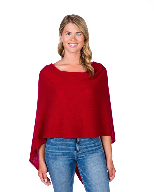 A smiling blonde woman wearing a bright red knit poncho-style top with blue jeans, standing against a plain white background.