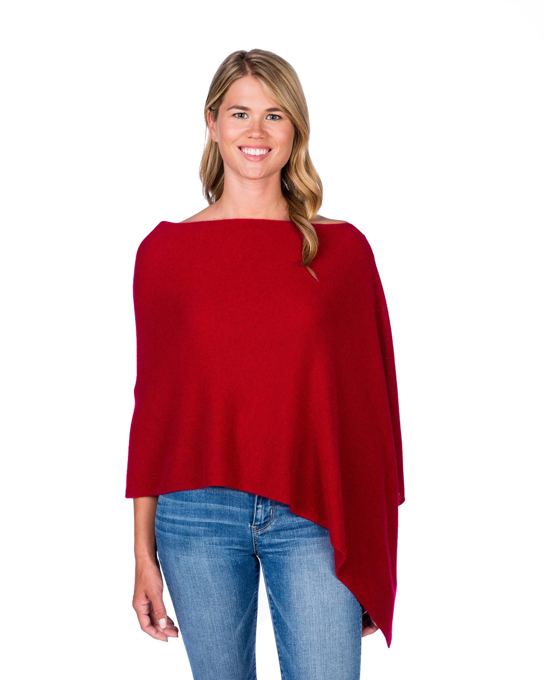 A woman modeling a red knit poncho top with an asymmetrical hem and wide boat neckline, paired with blue jeans, against a plain white background.