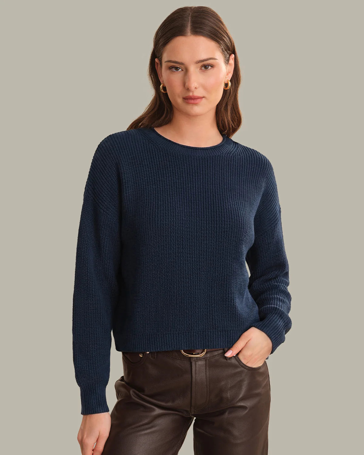 A woman modeling a navy blue ribbed knit sweater with long sleeves and a crew neck, paired with dark brown leather pants, against a solid light gray background.
