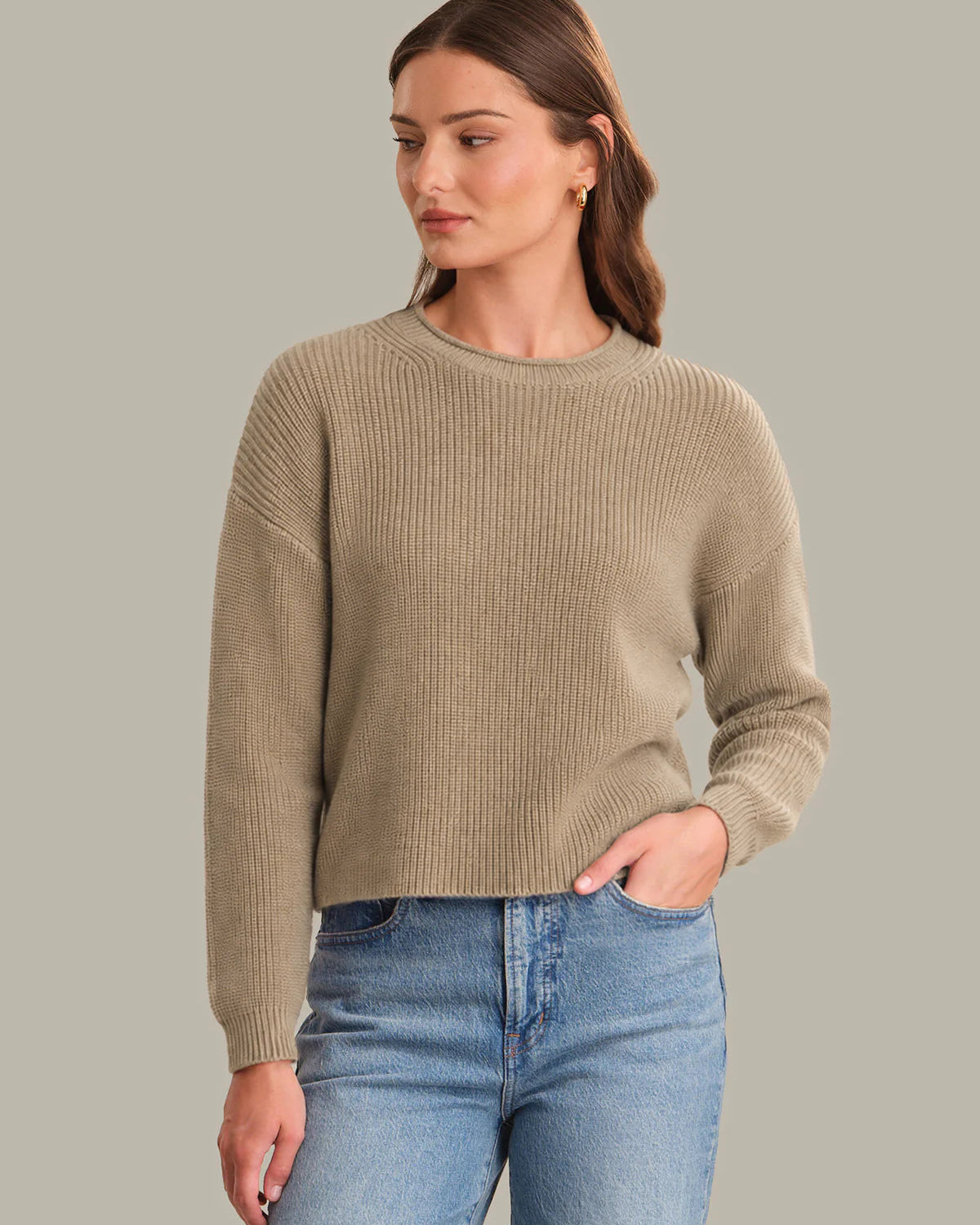 A woman wearing a beige ribbed knit sweater and light blue jeans, standing against a solid gray studio background.