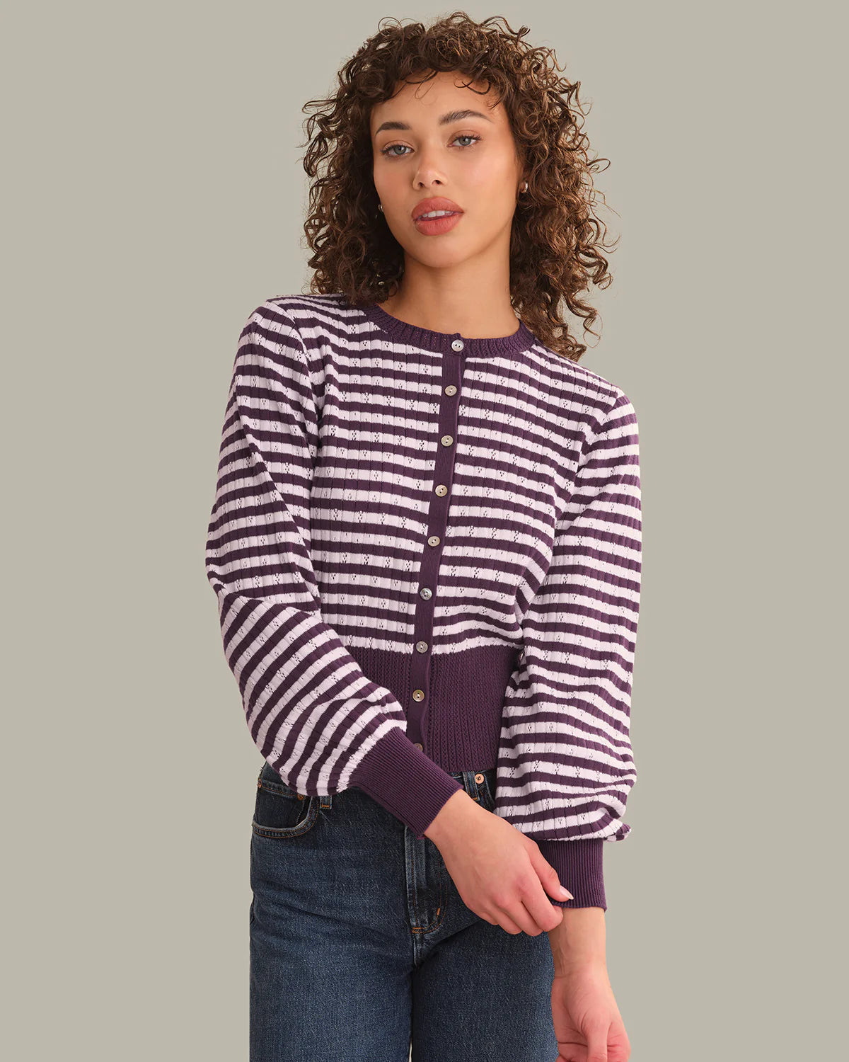 A woman with curly brown hair wearing a purple and white striped long-sleeve button-up cardigan with dark purple ribbed trim, paired with dark blue jeans, against a solid light gray background.