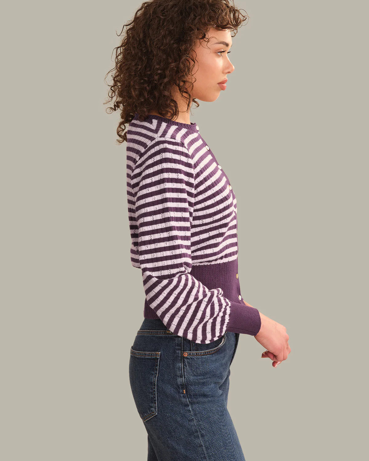 A woman shown in side profile wearing a purple and white horizontally striped long-sleeve knit top with a solid purple waistband, paired with blue jeans, against a solid gray studio background.