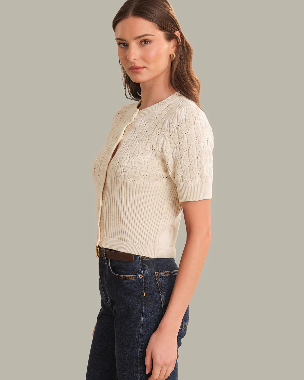 Woman wearing a cream knit cardigan over a ribbed top with dark jeans on a gray background