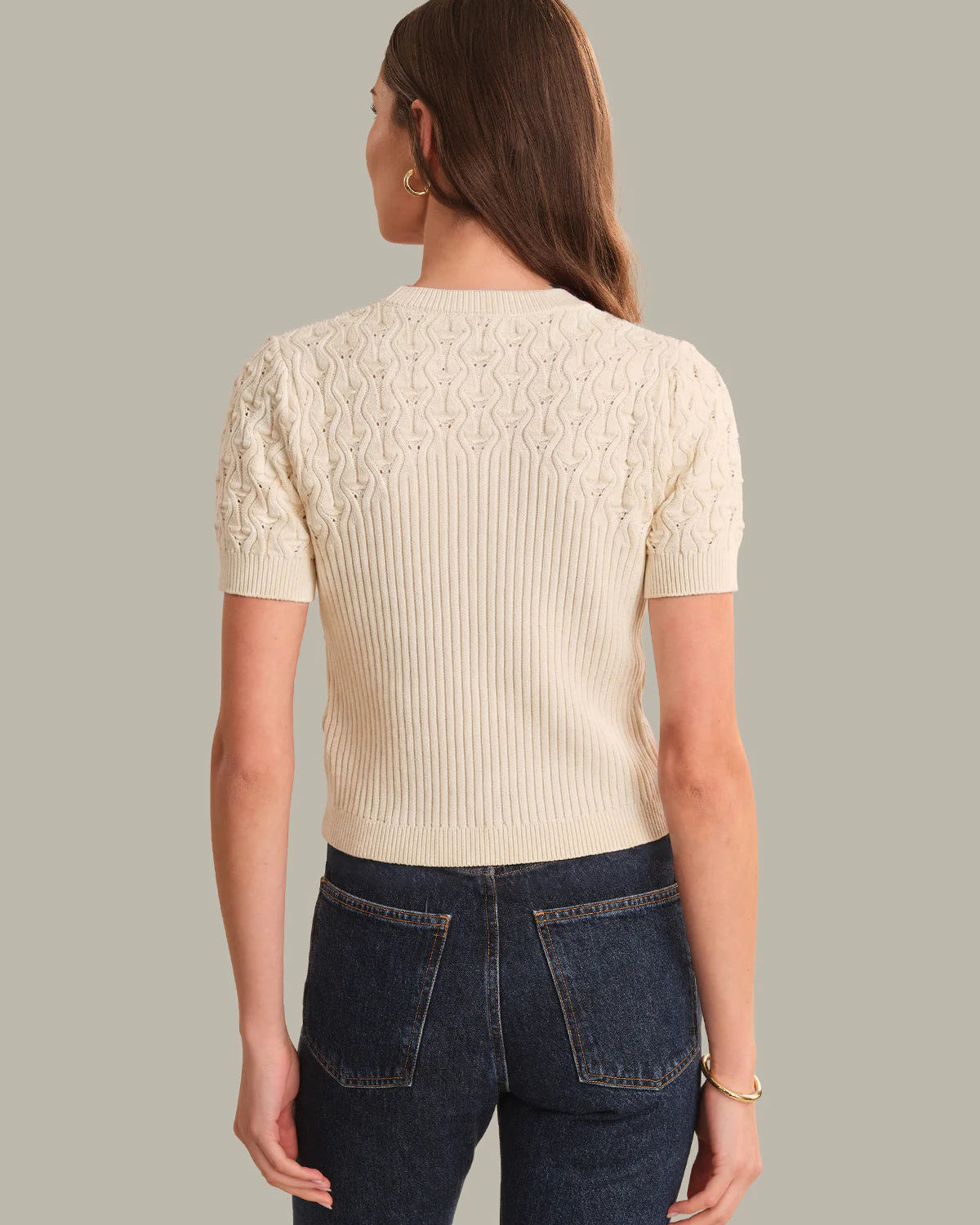 Woman wearing a cream ribbed sweater and dark blue jeans on a gray background