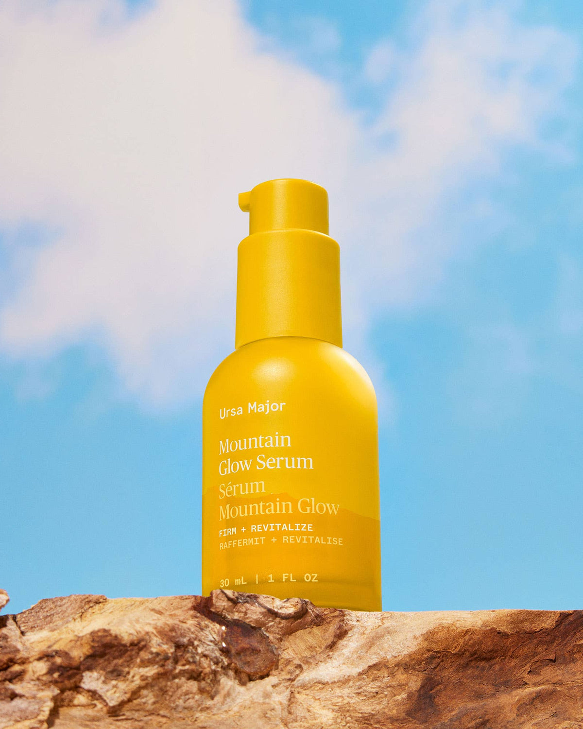Yellow serum bottle labeled 'Mountain Glow Serum' on a wooden surface with a blue sky background