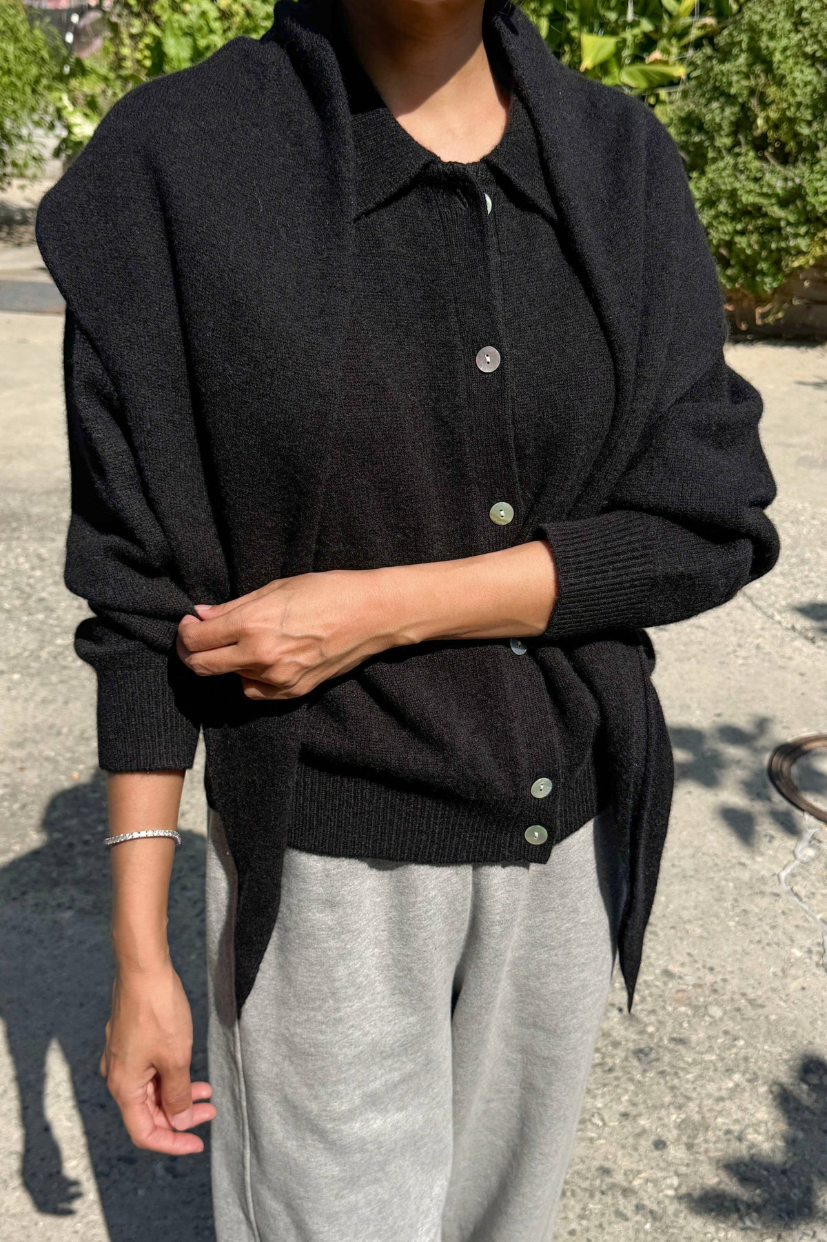 A person outdoors wearing a black knit cardigan with a collar and front buttons over a dark top, paired with light gray sweatpants.
