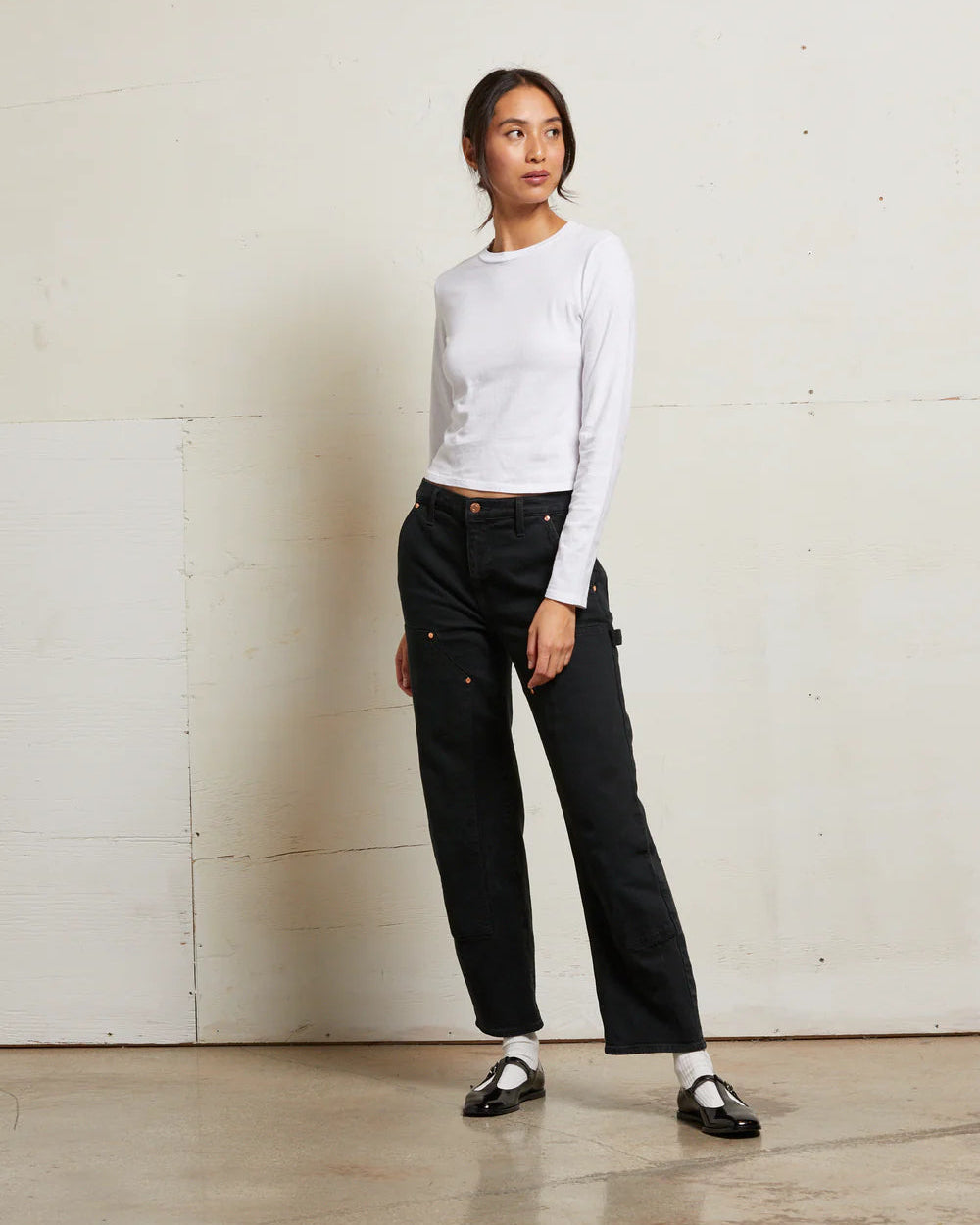 Woman wearing a white long-sleeve top and black pants against a plain wall.