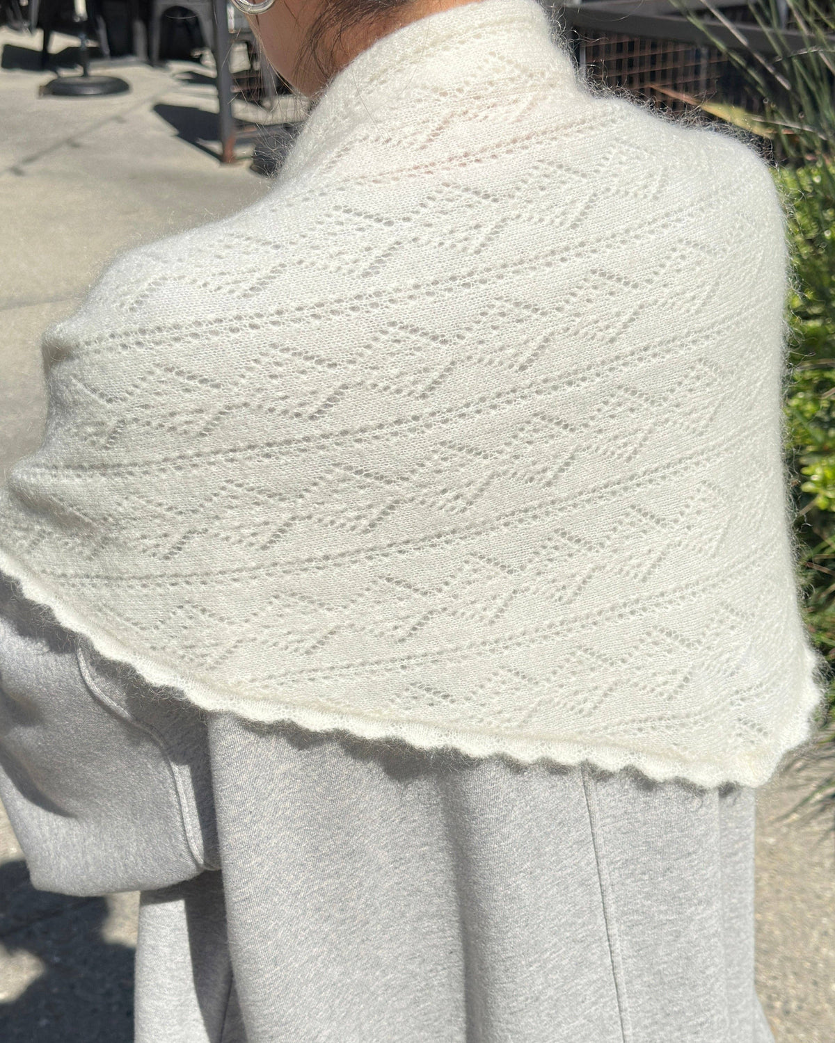 A close-up of a person outdoors wearing a cream-colored knit shawl with a geometric diamond pattern and a scalloped edge over a light gray top.