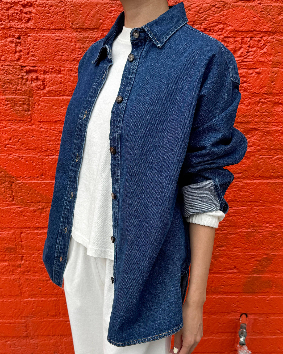 A person wearing an oversized dark blue denim button-up shirt over a white top and white pants, standing in front of a bright orange-red brick wall.