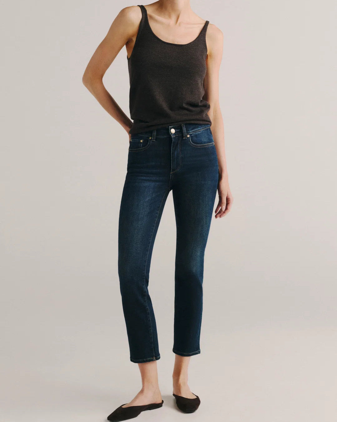 A model wearing dark blue cropped jeans, a black tank top, and black flat shoes, standing against a light gray studio background.