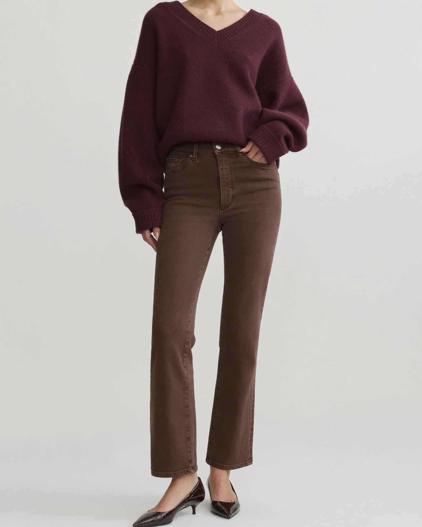 A model wearing a dark burgundy v-neck knit sweater and brown straight-leg jeans, paired with black pointed-toe heels, against a solid light gray studio background.