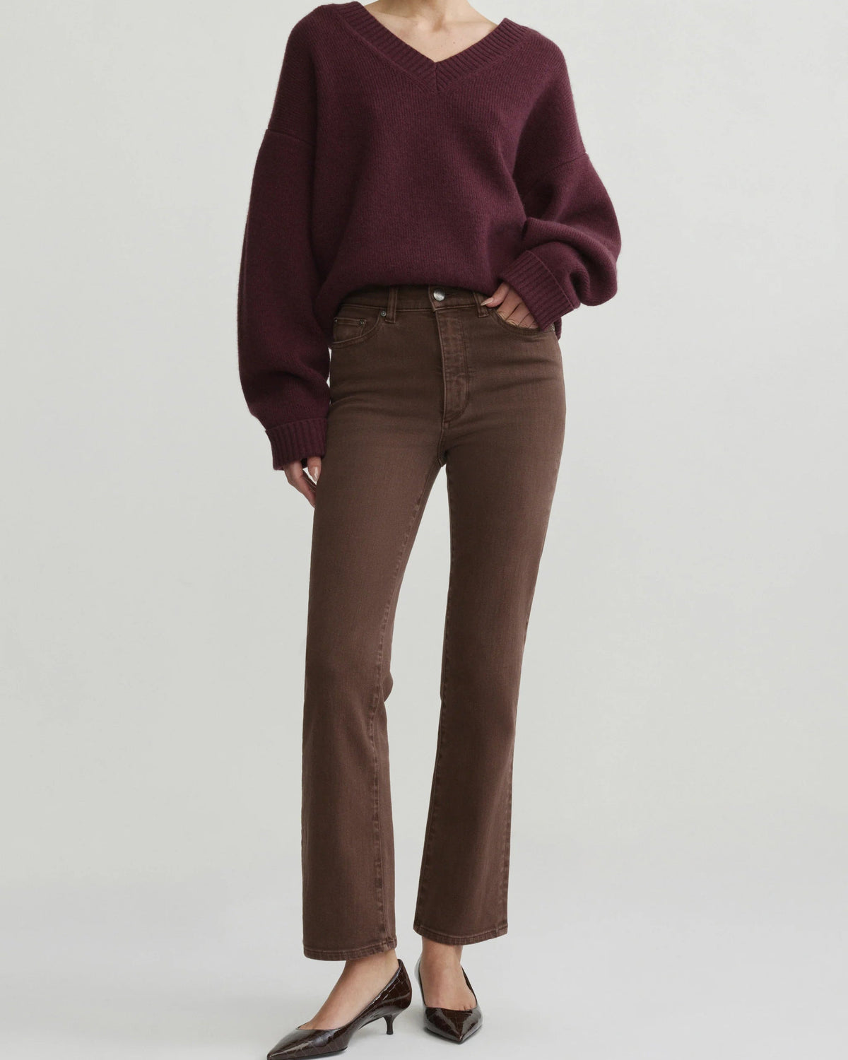 A model wearing a dark burgundy v-neck knit sweater and brown straight-leg jeans, paired with black pointed-toe heels, against a solid light gray studio background.