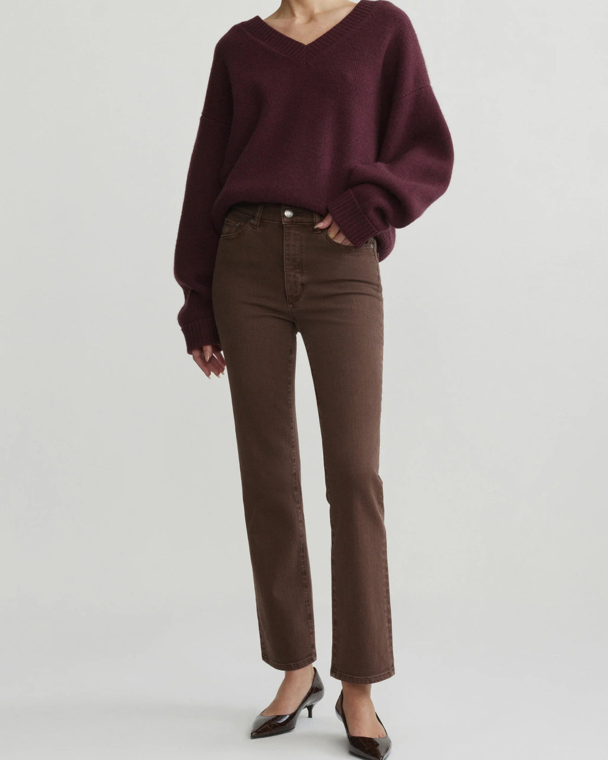 A model wearing a dark burgundy knit V-neck sweater, brown straight-leg jeans, and black pointed-toe heels, standing against a light gray studio background.