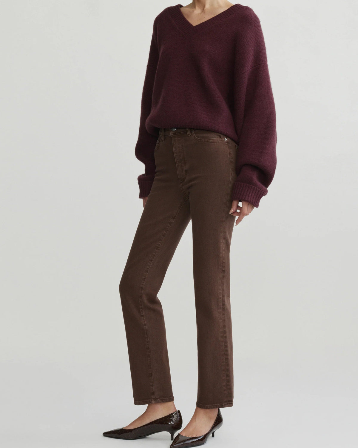 A model wearing a dark burgundy V-neck knit sweater, brown straight-leg jeans, and black pointed-toe flats, photographed against a light gray studio background.
