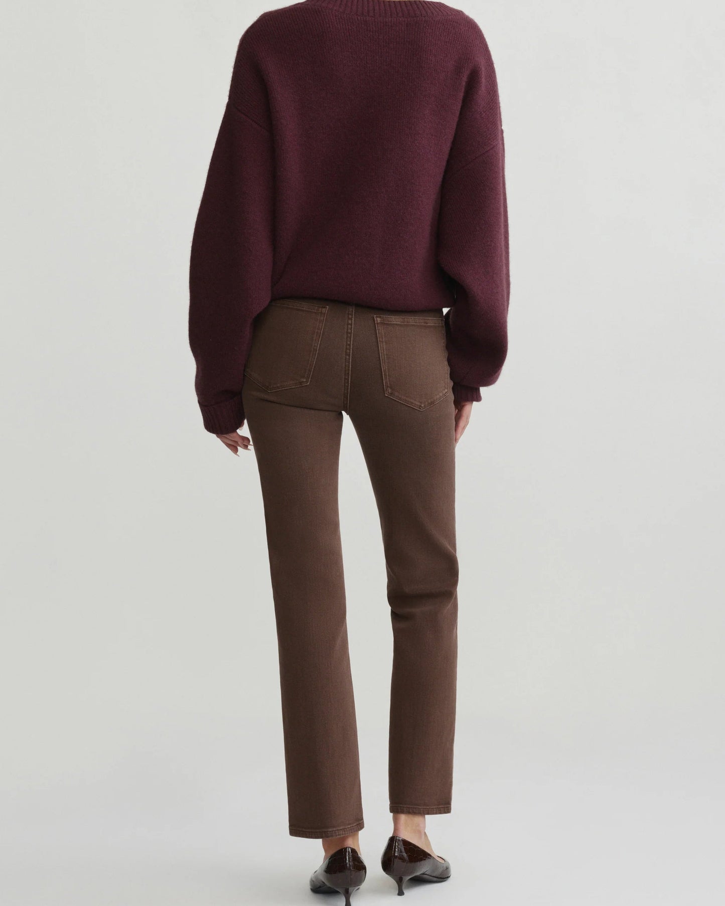 A model shown from the back wearing a dark burgundy knit sweater, brown straight-leg jeans, and dark brown pointed-toe flats against a light gray studio background.