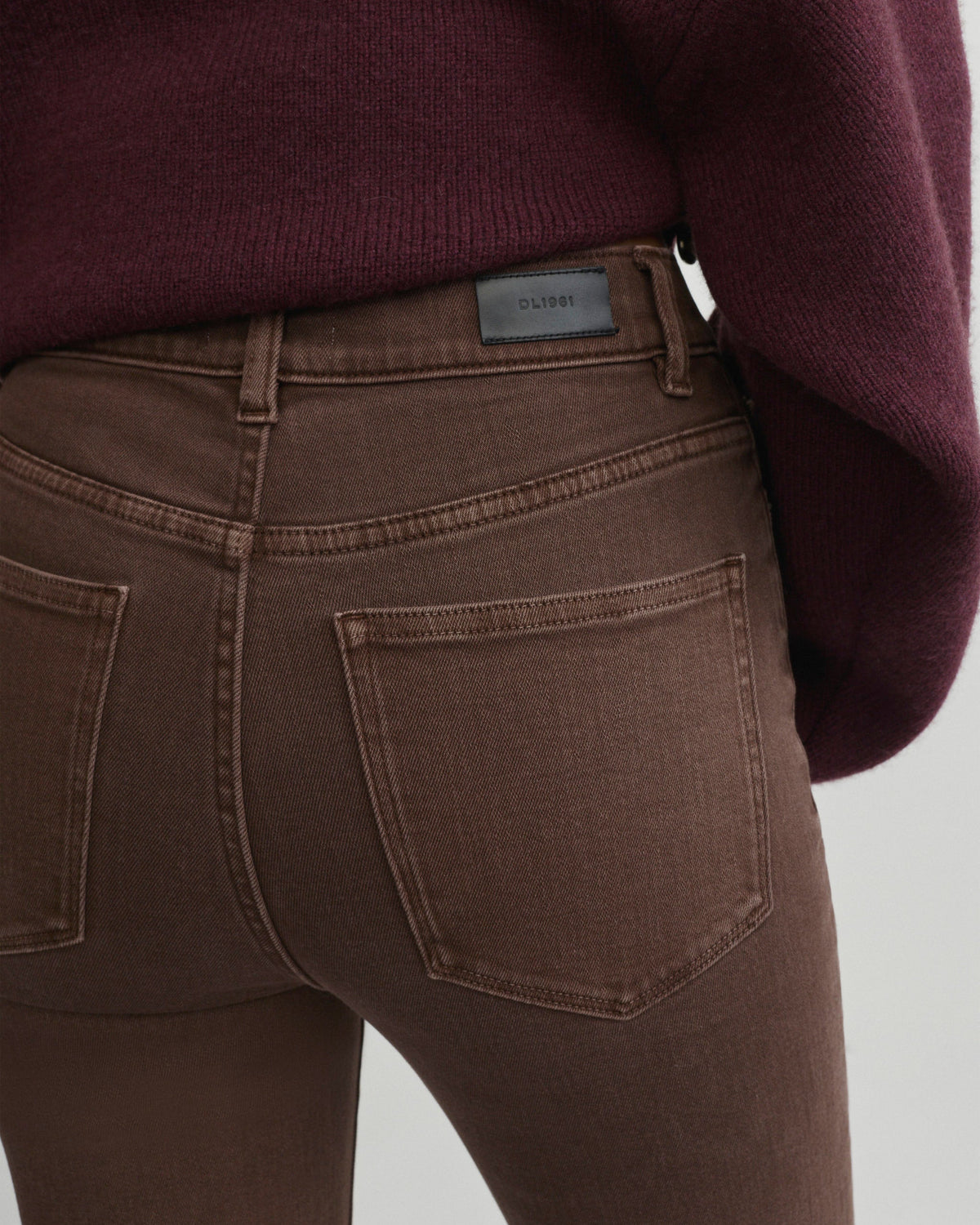 Close-up back view of a person wearing dark brown skinny jeans and a burgundy sweater, showing the waistband, belt loop, and back pocket details.