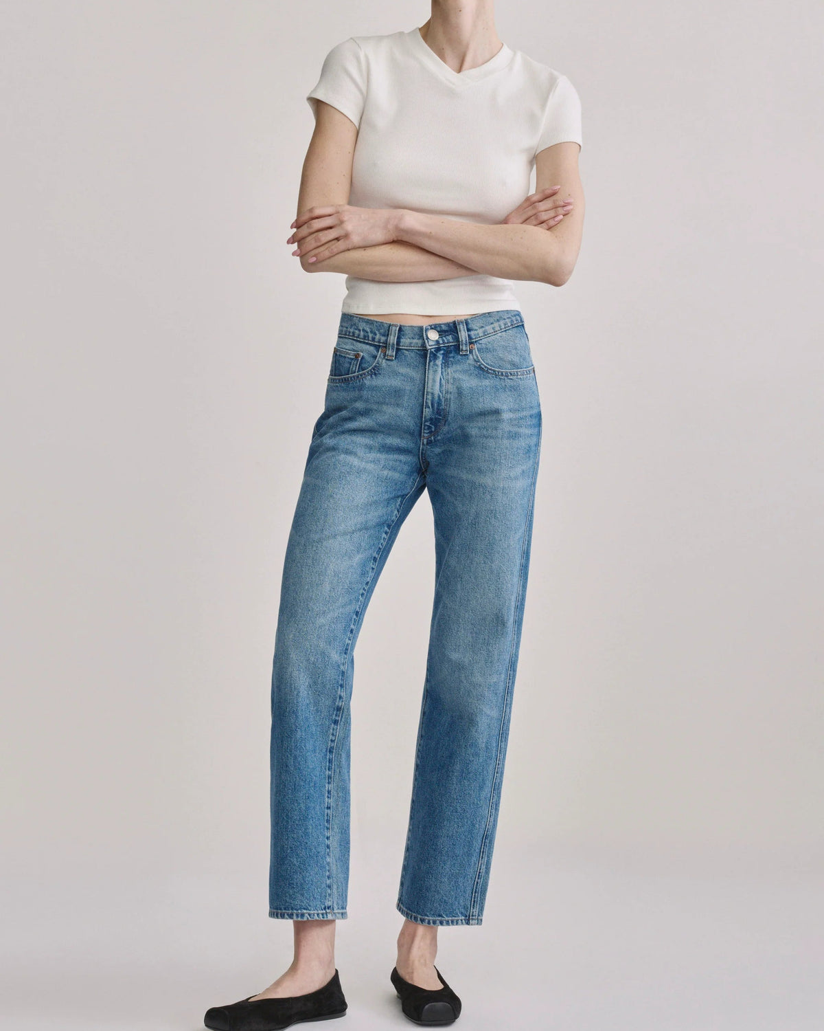 A model wearing high-waisted, straight-leg blue jeans with a faded wash, styled with a white short-sleeve crop top and black flats, against a light gray studio background.