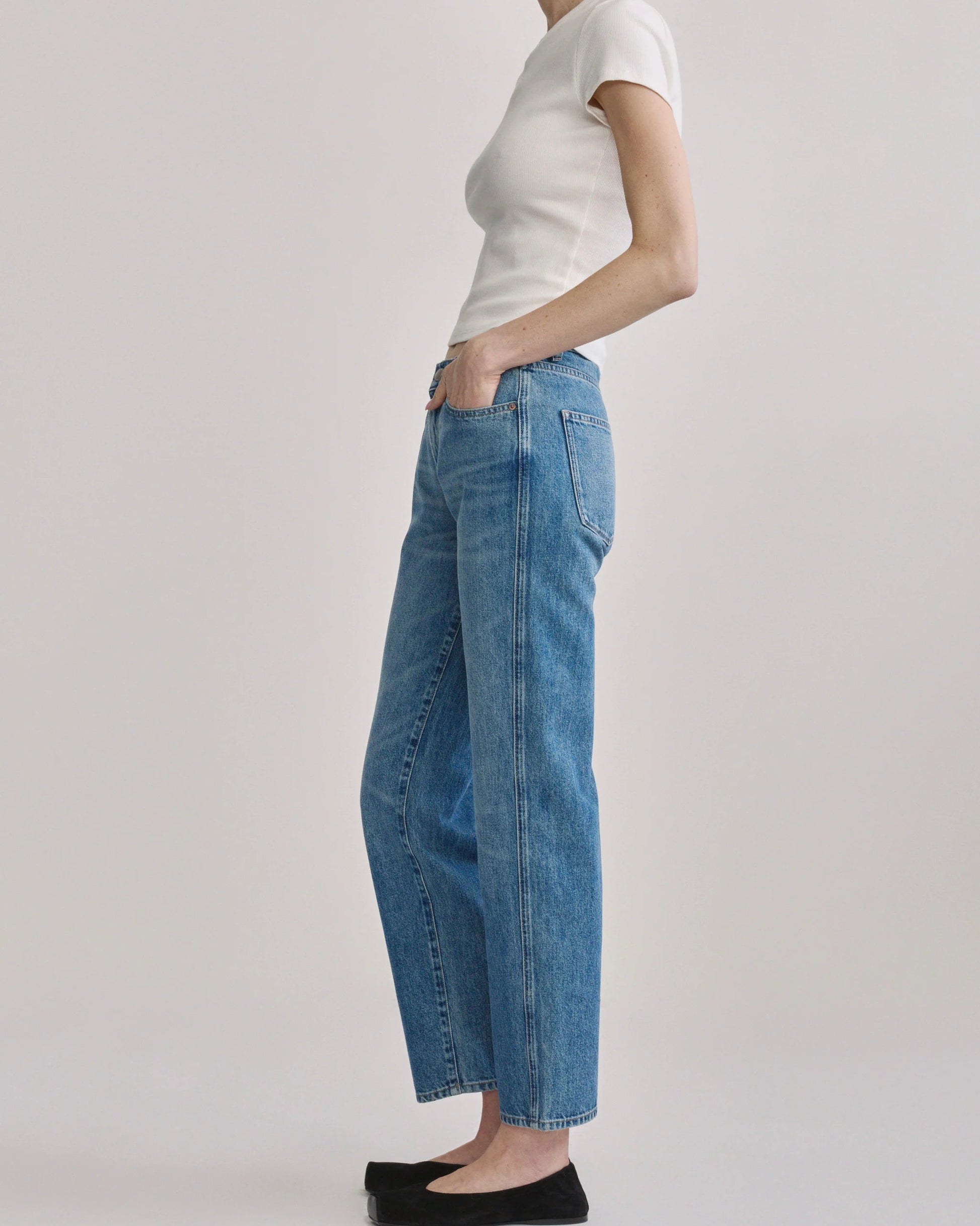 A model shown from the side wearing light-wash blue wide-leg cropped jeans, a white short-sleeve top, and black flats against a plain light gray studio background.