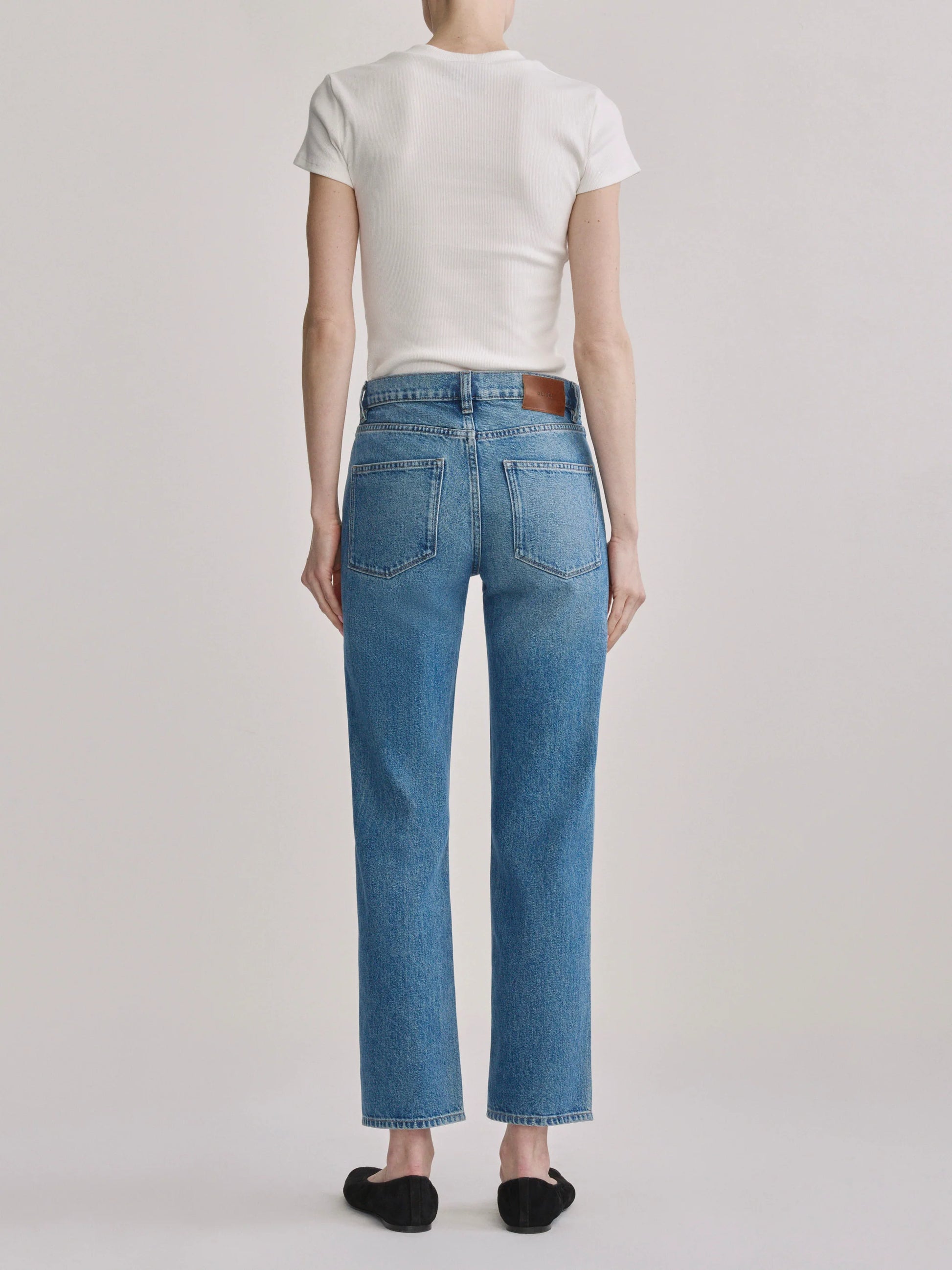 A model shown from the back wearing light-wash blue straight-leg jeans, a white t-shirt, and black flat shoes against a light gray studio background.