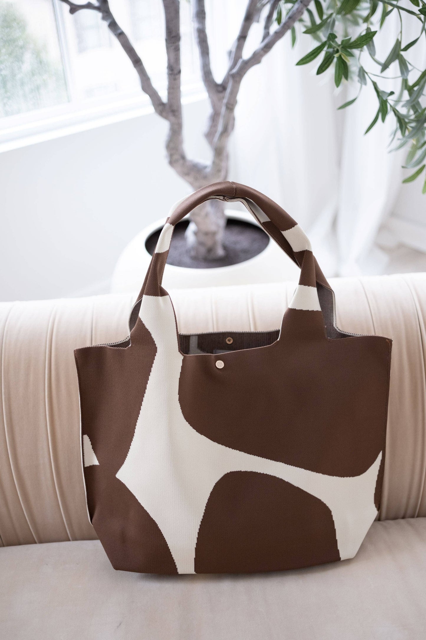 Brown and white patterned tote bag on a light-colored surface with a plant in the background