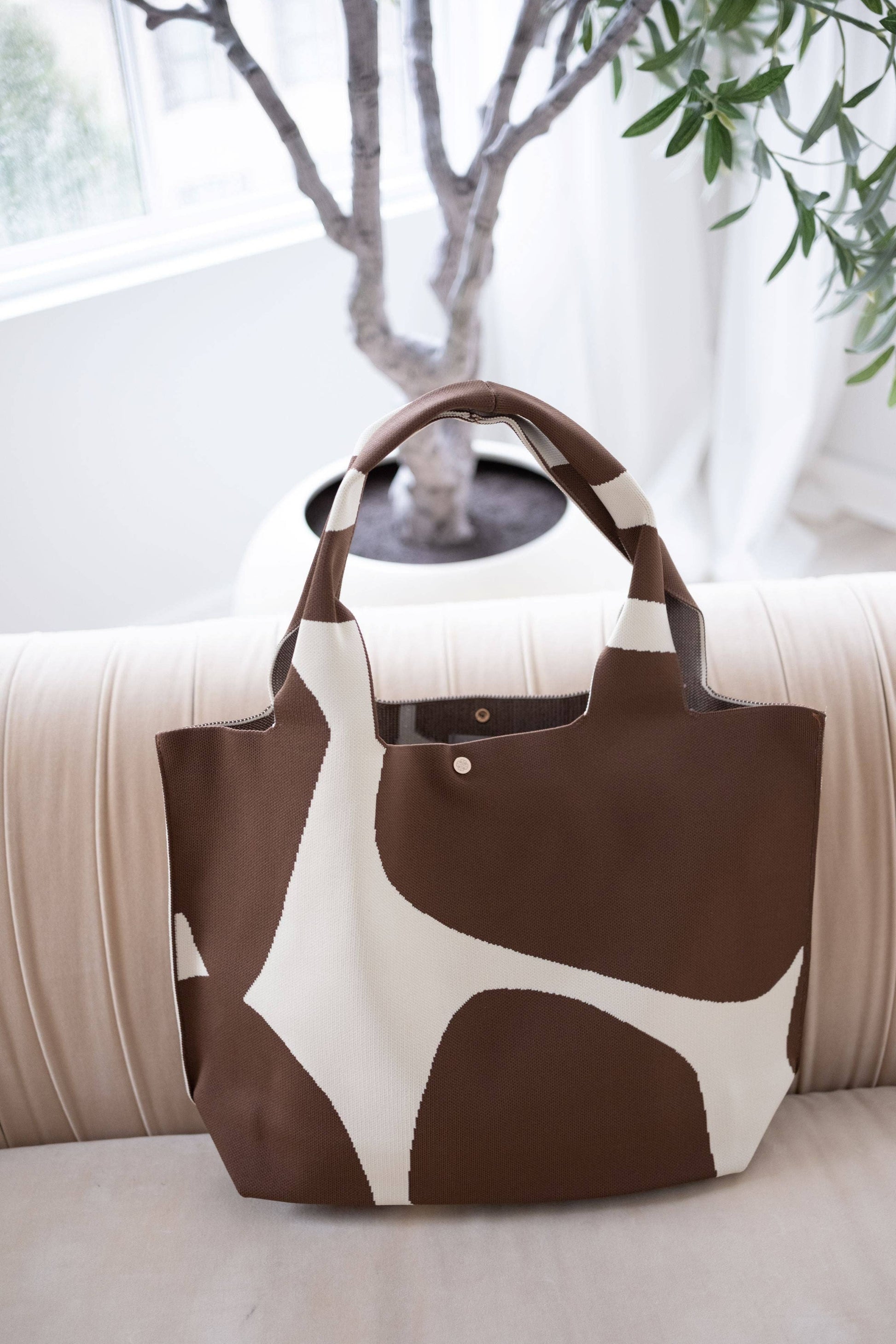 Brown and white patterned tote bag on a light-colored surface with a plant in the background