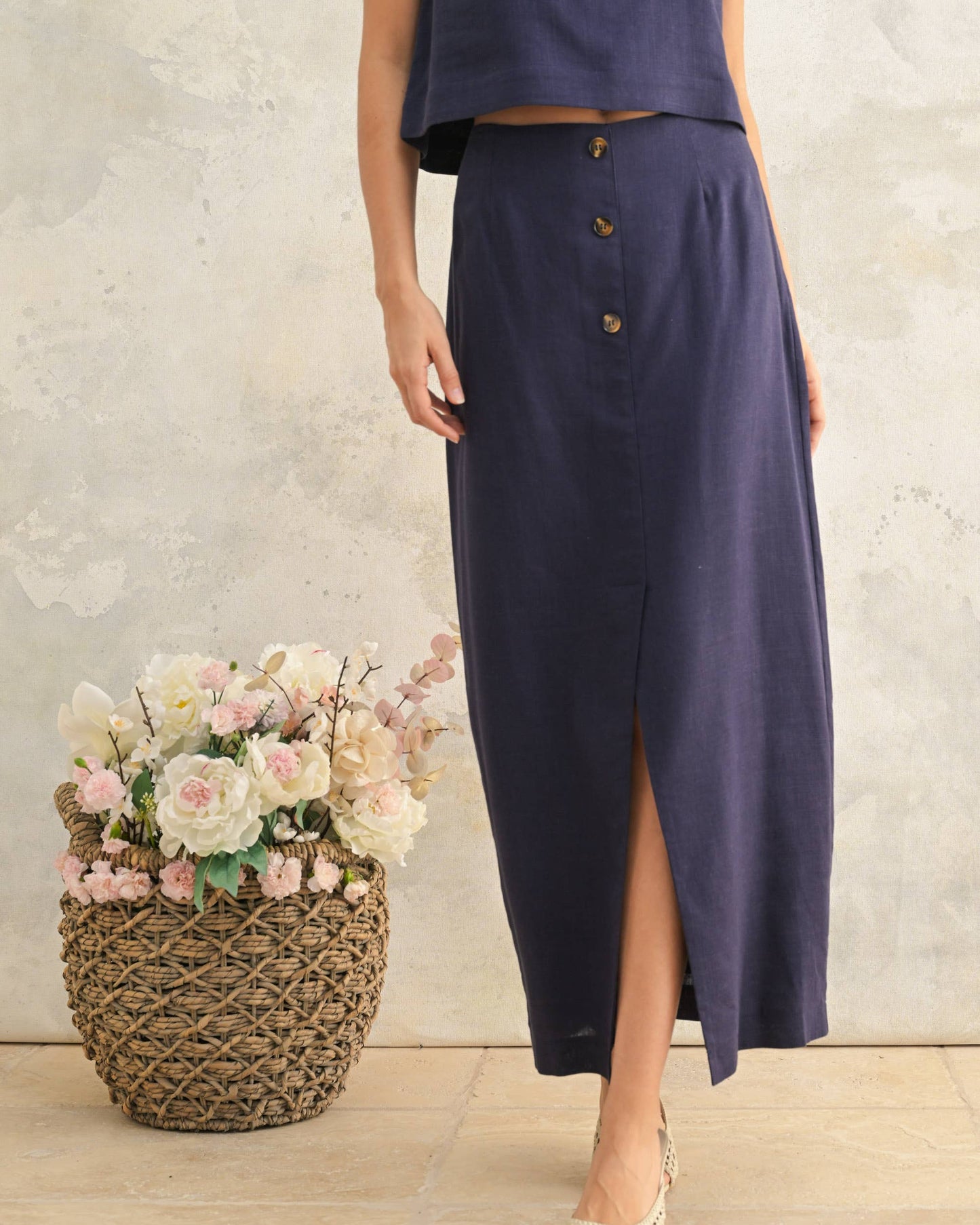 Woman wearing a long navy skirt with a textured wall and flowers in the background