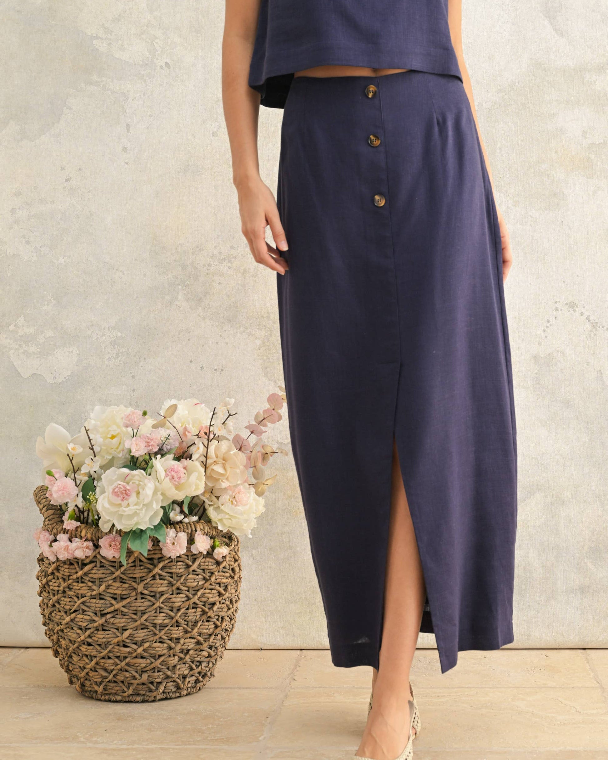 Woman wearing a long navy skirt with a textured wall and flowers in the background