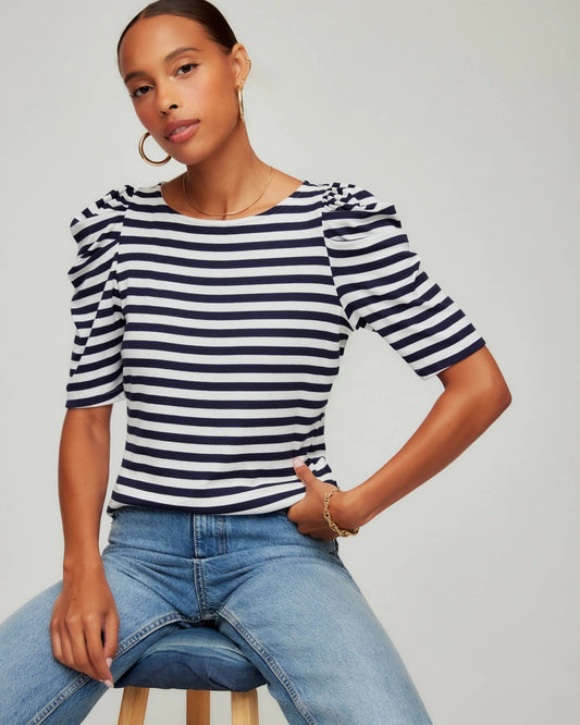 Woman wearing a navy and white striped shirt with ruffled sleeves and blue jeans on a gray background