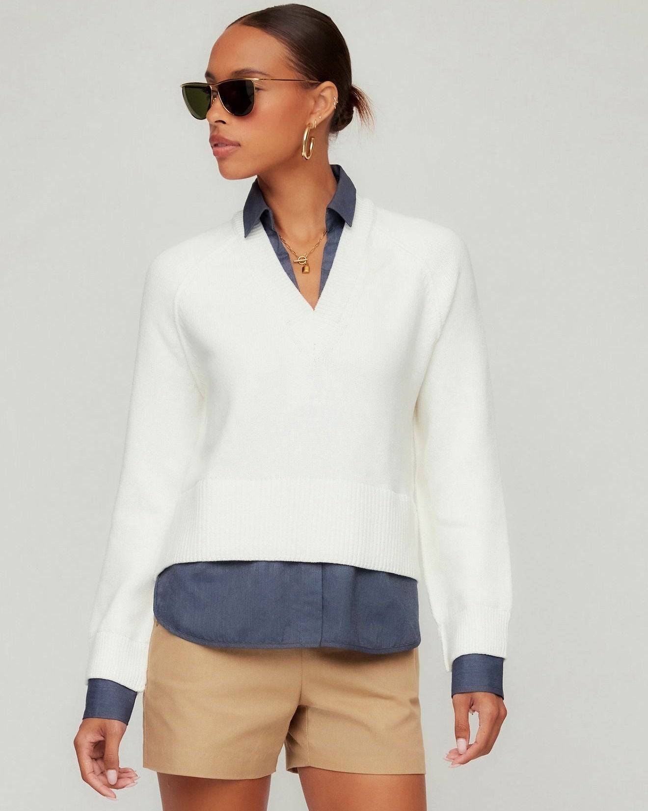Woman wearing a white sweater with blue accents and beige shorts on a gray background