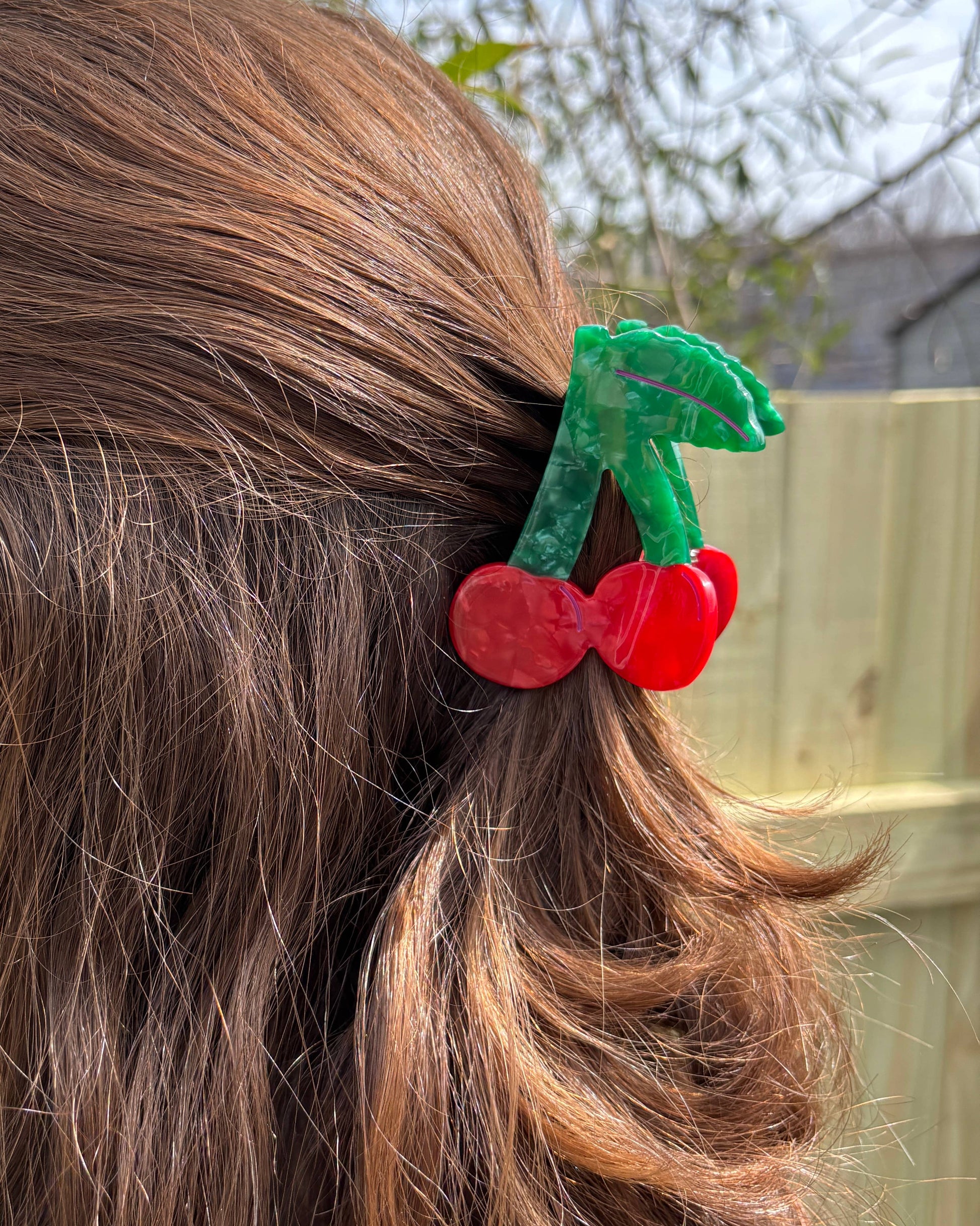 Close up of a cherry-shaped hair clip on a person's hair with a blurred outdoor background