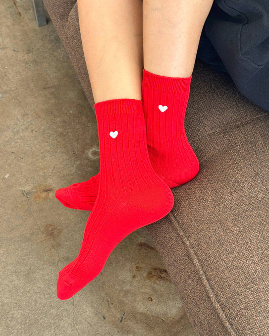 Red socks with heart designs worn by a person sitting on a concrete surface.
