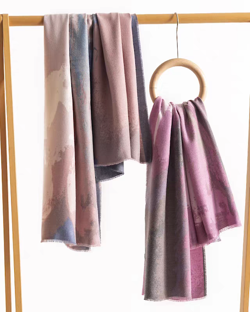 Two lightweight patterned scarves in muted dusty pink, mauve, and gray tones hang from a wooden clothing rack against a white background.
