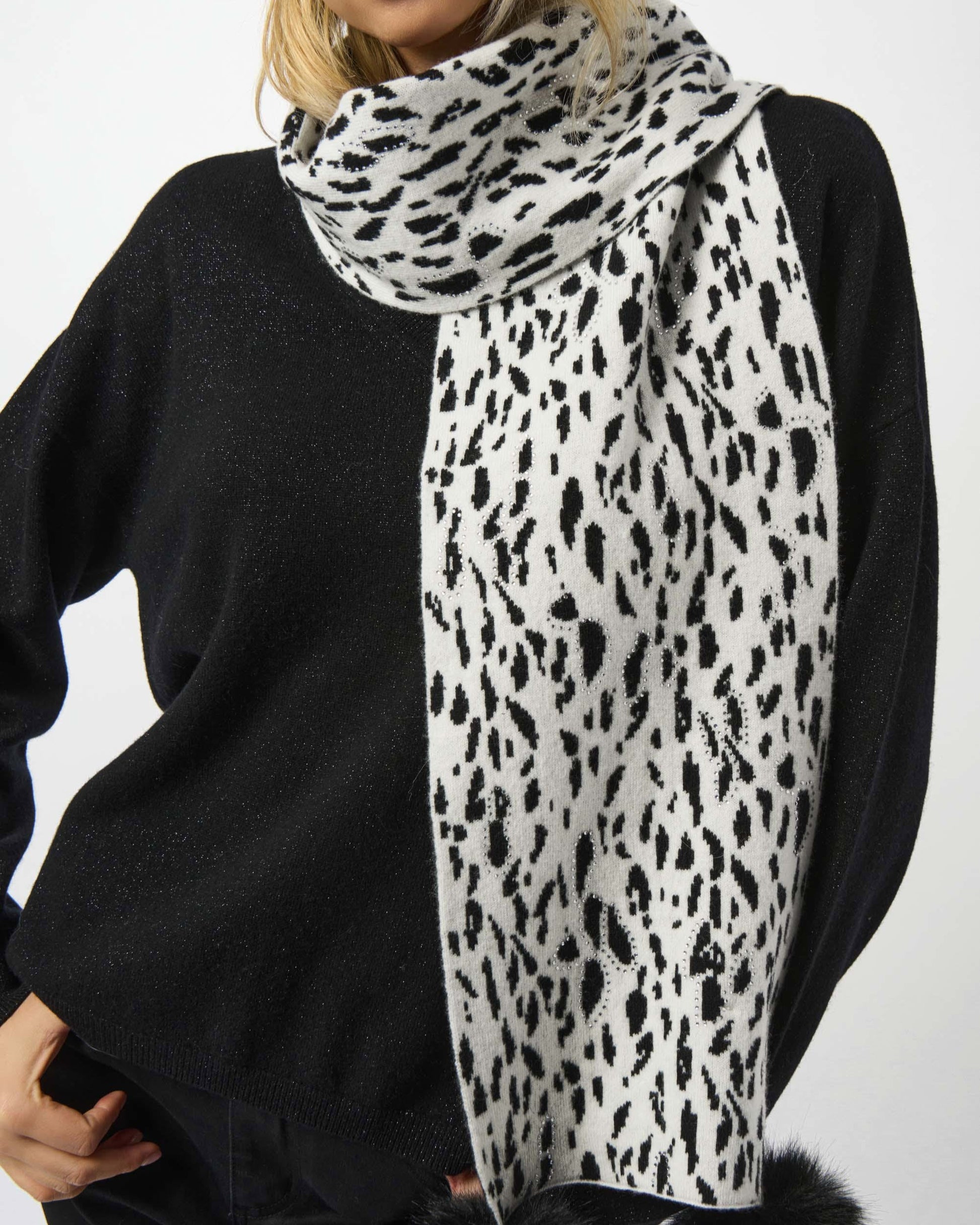 Person wearing a black sweater and a white scarf with black leopard print and pom-poms on a white background